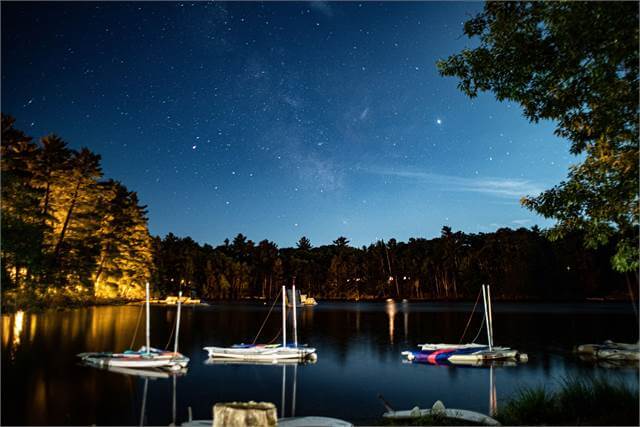Sailboats are docked on a calm lake at night, surrounded by trees. The sky is clear, filled with stars, and the water reflects the night scenery. Some trees are softly lit along the shoreline.