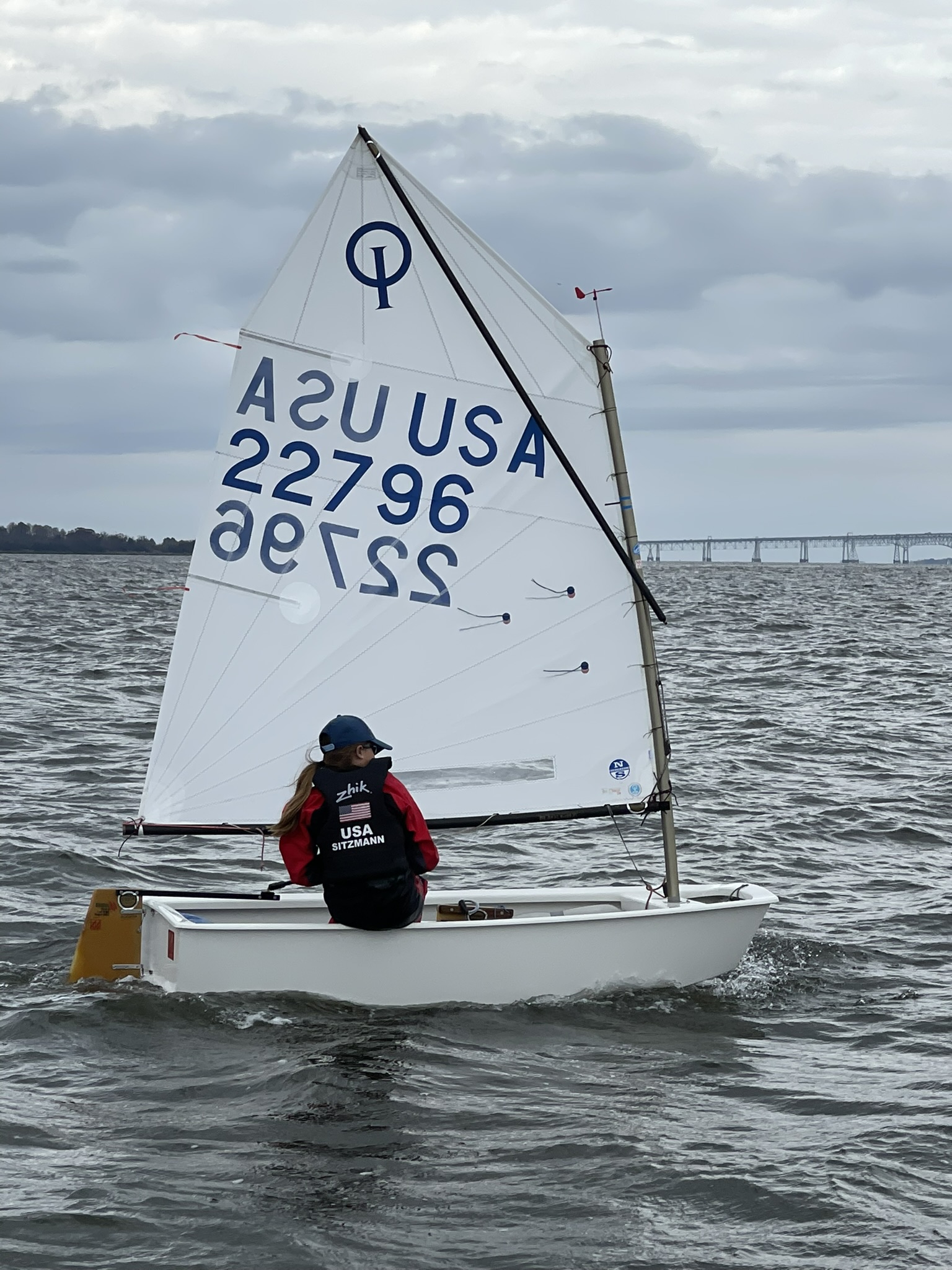 A person wearing a life jacket and cap is sailing a small white dinghy with the sail number 22796 on a cloudy day, with a bridge visible in the distance over choppy water.