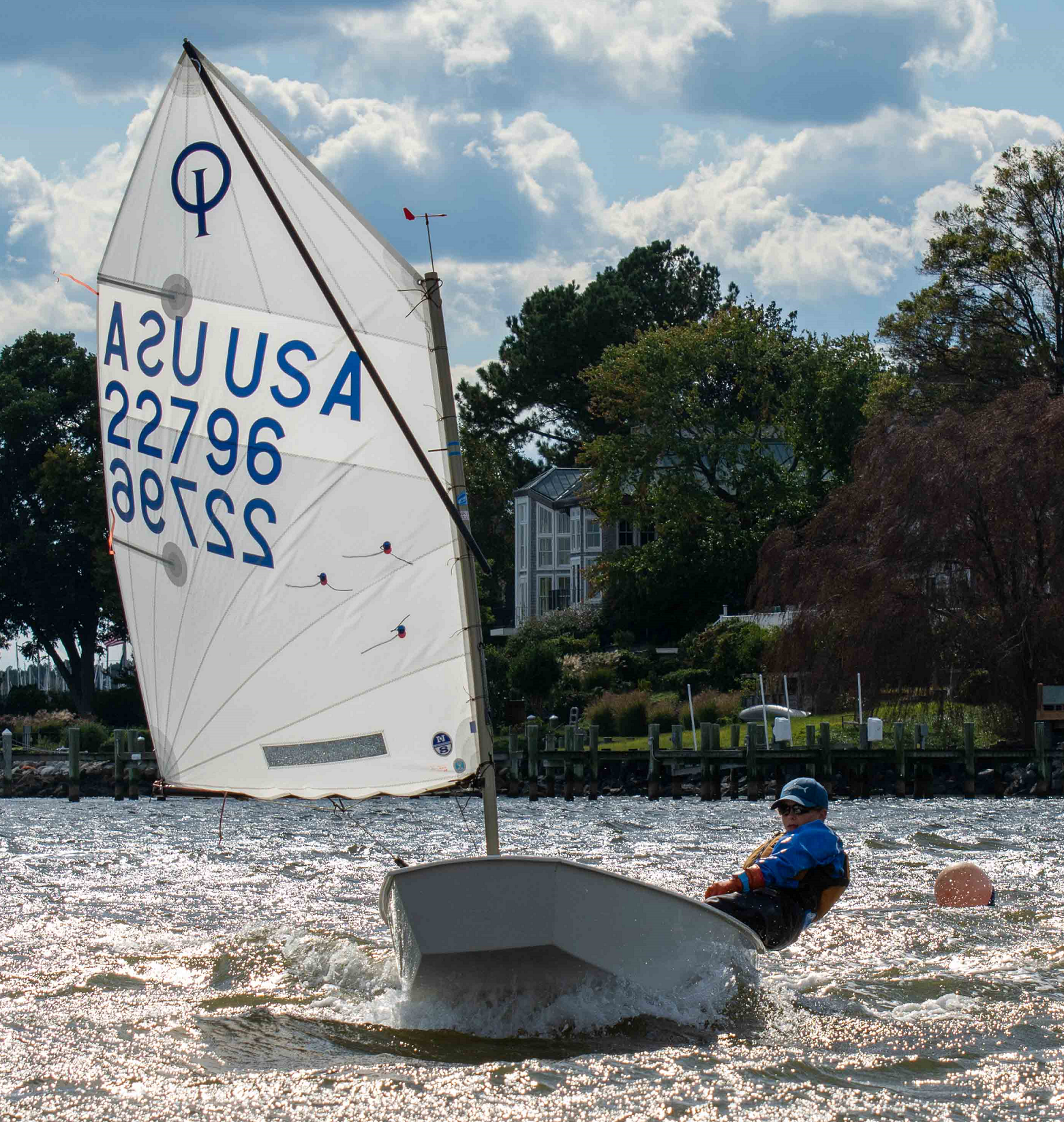 A person wearing a helmet and life jacket sails a small white boat with a triangular sail marked "USA 22796" on a sunlit body of water, with houses, trees, and a dock in the background.