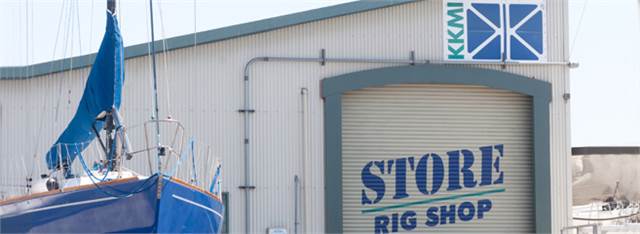 A blue sailboat is positioned beside a building with a large sign reading 'STORE RIG SHOP.' The building also displays a KKMI logo above a rolling door.