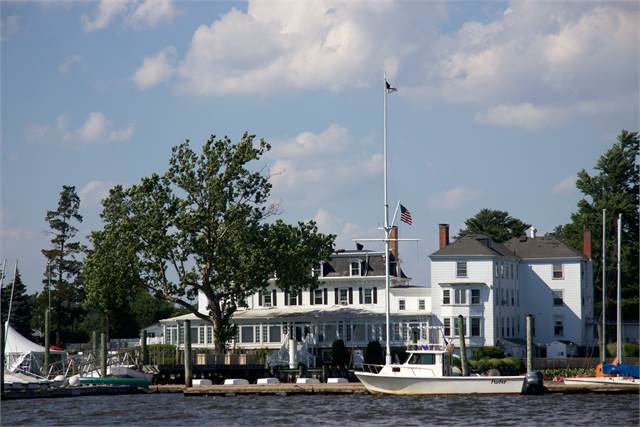 A waterfront yacht club with white buildings, sailboats docked along the shoreline, a large tree, and an American flag on a tall flagpole, under a partly cloudy sky.
