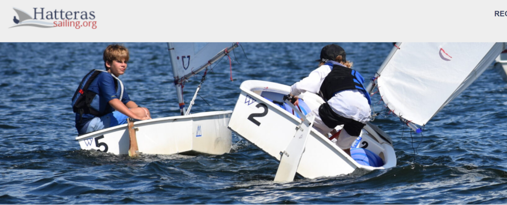 Two children in life jackets sail small white boats on a sunny day, with the Hatteras Sailing logo in the top left corner of the image. One child looks toward the camera while the other maneuvers their boat.