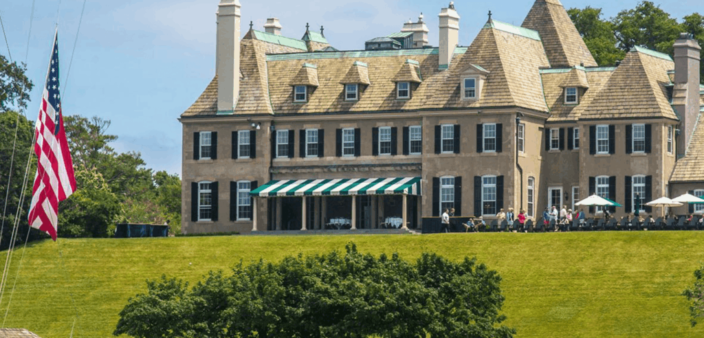 A large, historic mansion with tan walls and a green roof sits atop a grassy hill. An American flag flies on the left, and people gather on the patio under a green-and-white striped awning. Trees surround the property.
