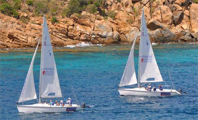 Two white sailboats with “Offshore Sailing School” logos glide on clear blue water near a rocky, coastal shoreline. Each boat has several people onboard, enjoying the sunny day.