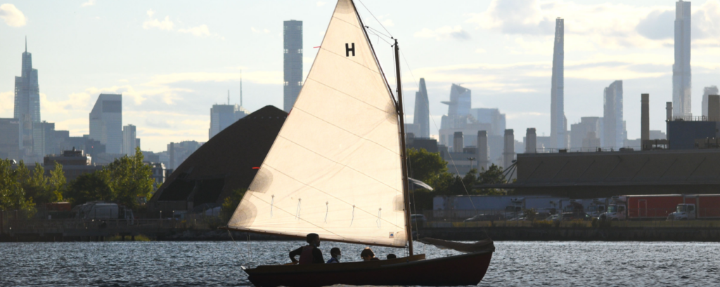 A small sailboat with four people glides on the water in front of a city skyline, with tall buildings and a clear sky in the background. The sunlight illuminates the boat’s sail.