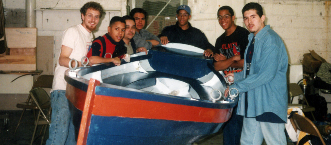 Six young men stand around a small painted rowboat inside a workshop, smiling at the camera. The boat is blue with a red stripe. The background shows tools, chairs, and a cinderblock wall.