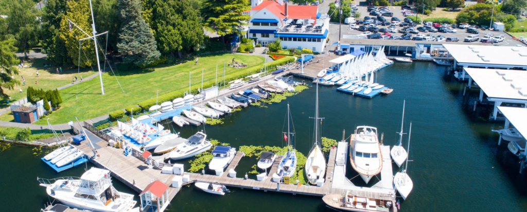 Aerial view of a marina with several boats docked along wooden piers, surrounded by green lawns and trees. Buildings and a parking lot with many cars are visible in the background.
