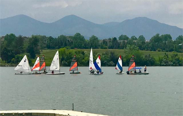 Several small sailboats with colorful sails glide on a lake, with people onboard. Lush green trees and hills line the shore, and blue mountains rise in the background under a cloudy sky.