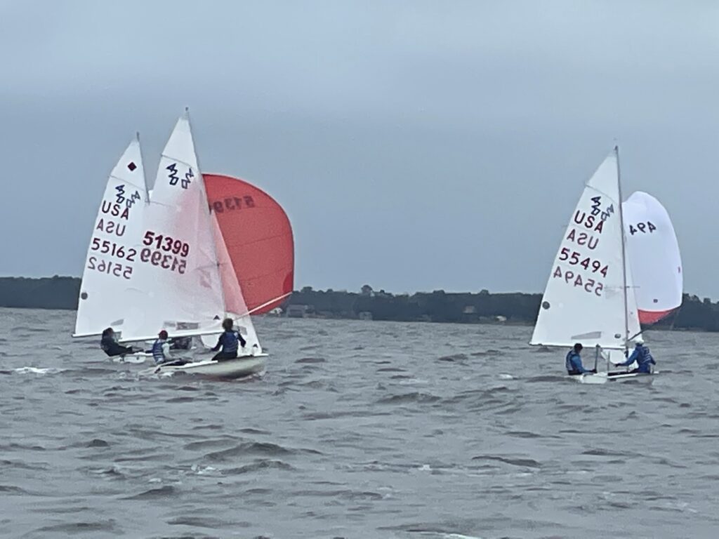 Three small sailboats with red and white sails race on choppy water under a cloudy sky. Four sailors are visible, each controlling a boat, and a shoreline with trees is seen in the background.