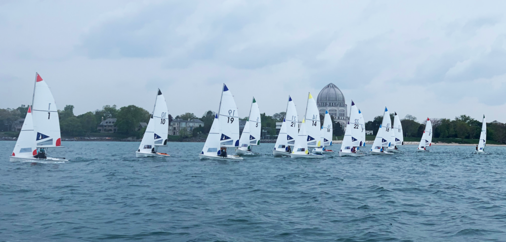 A group of small sailboats with numbered sails race on a lake under a cloudy sky, with trees and a large domed building visible on the shore in the background.