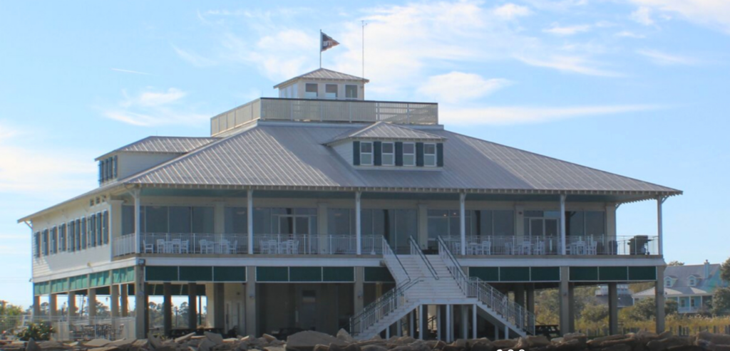 A large, white, two-story beachfront building with a metal roof, wraparound porch, and prominent central staircase, elevated on stilts with a flag flying on top. The sky is clear and blue.