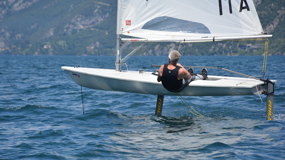 A person with gray hair steers a small sailboat equipped with hydrofoils, lifting above the blue water on a sunny day, with green hills and trees in the background.
