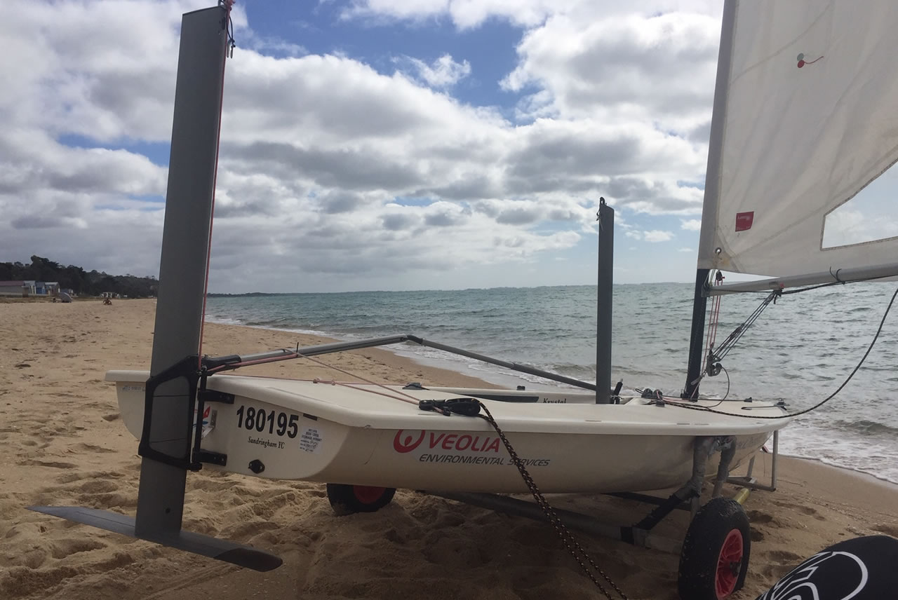 A small white sailboat with the number 180195 and "VEOLIA Environmental Services" on the side is on a sandy beach near the ocean, with its rudder and sail visible. The sky is cloudy.