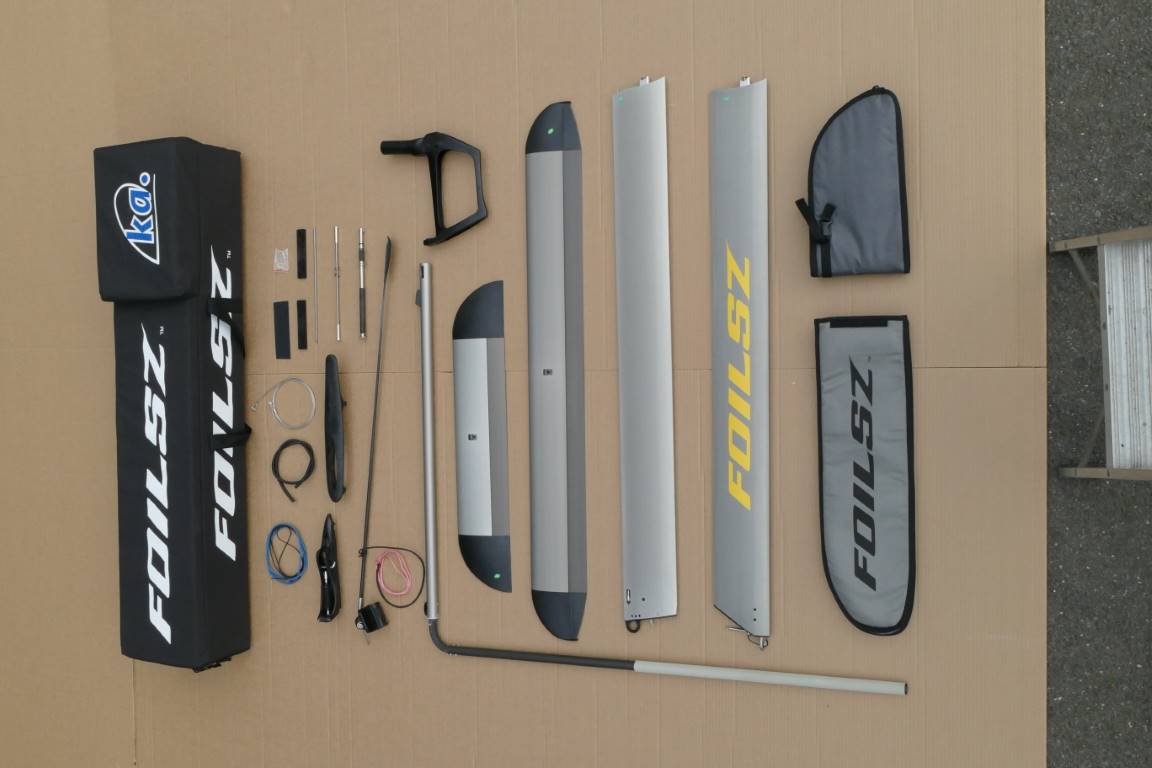 A foil kit for a windsurfing board is laid out on a cardboard surface. The kit includes metal foil wings, a mast, covers, screws, cables, tools, straps, and carrying bags labeled "FOILSZ" and "KA.