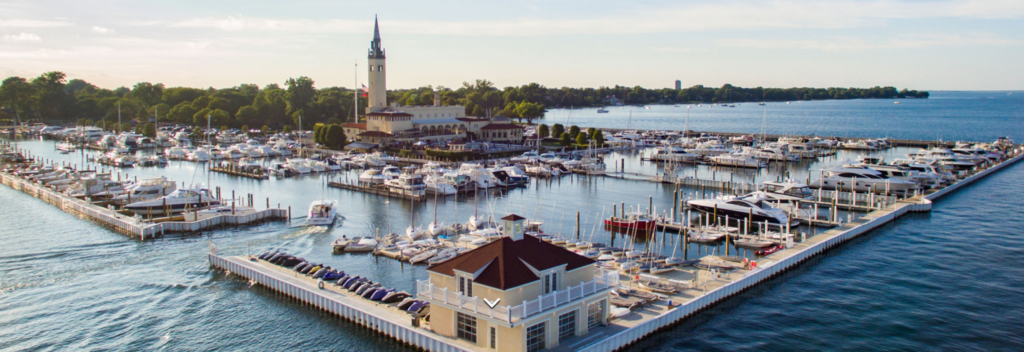 A marina filled with numerous boats and yachts, surrounded by docks and calm water, with a large building and clock tower in the background and trees lining the shore.