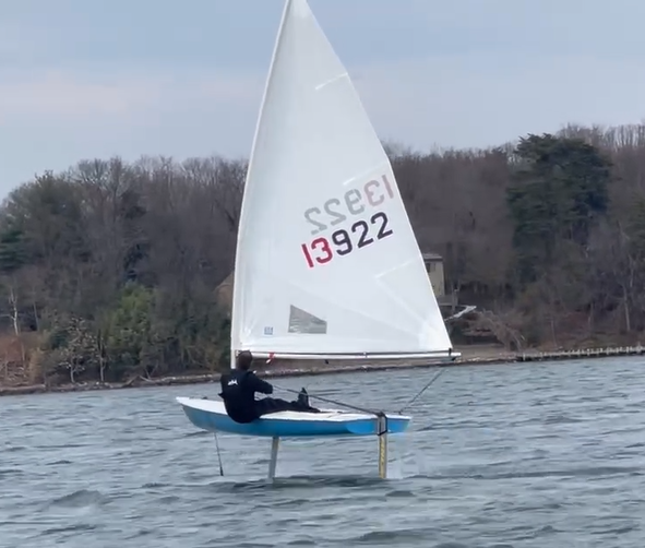 A person sails a small blue and white boat with the number 13922 on the sail. The boat is lifted above the water on hydrofoils, with trees and a shoreline in the background.