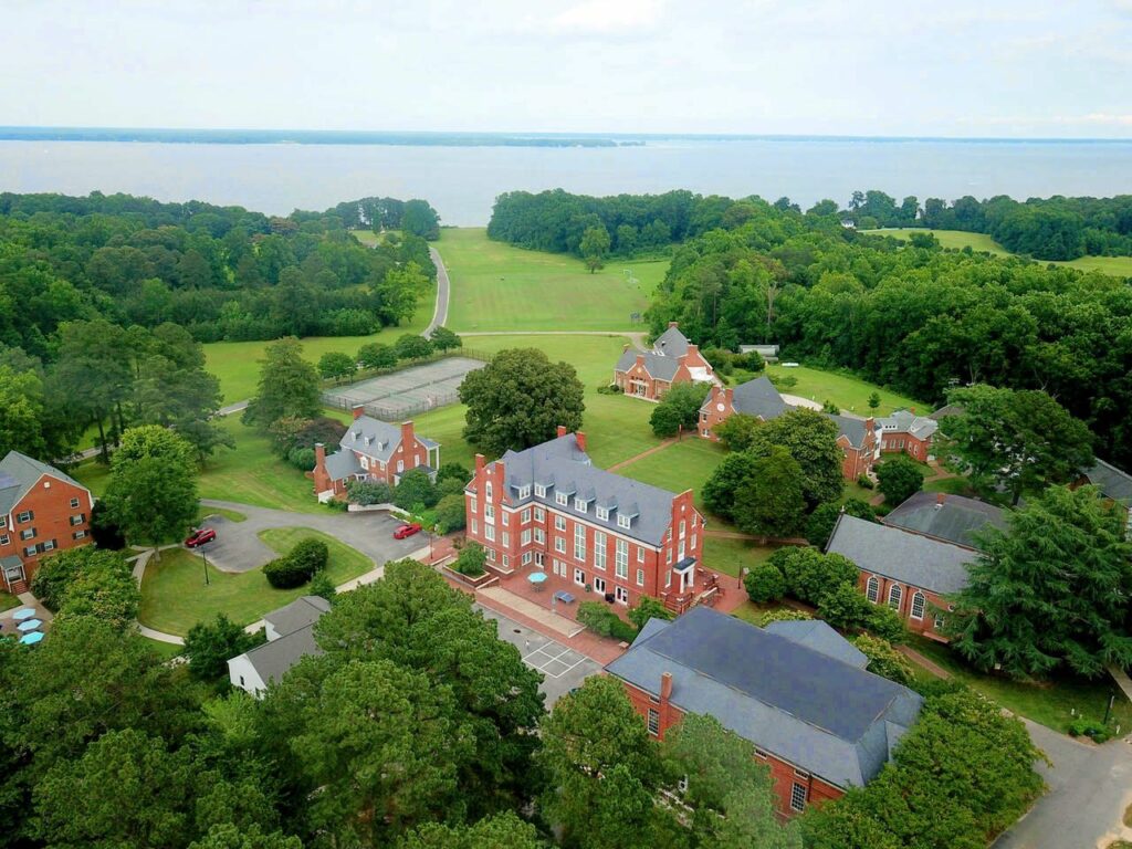 Aerial view of a scenic campus with red-brick buildings surrounded by green trees and lawns, tennis courts, and a road leading to a body of water in the background under a partly cloudy sky.