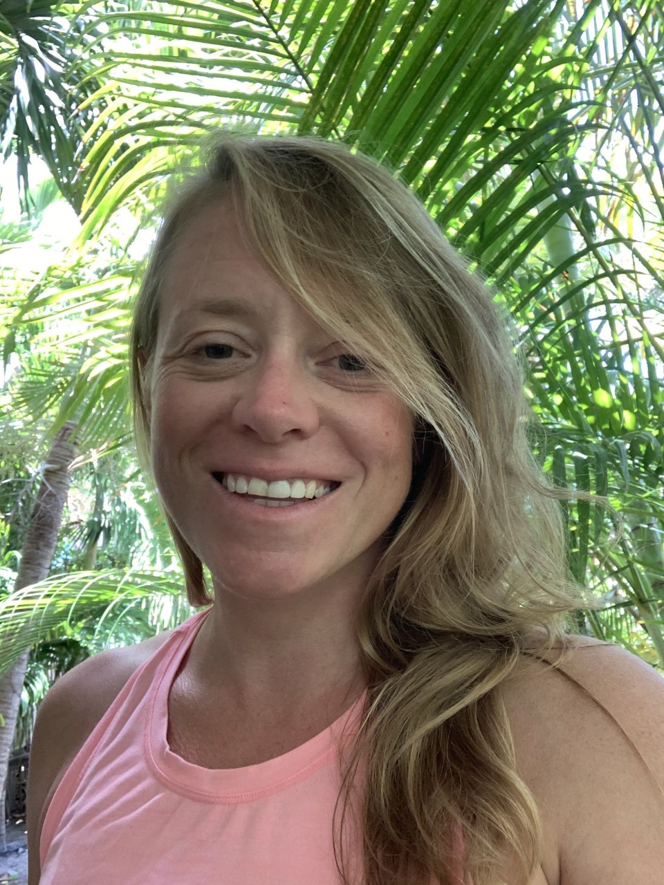 A woman with long blonde hair and a pink sleeveless top smiles at the camera, standing outdoors in front of lush green tropical plants.