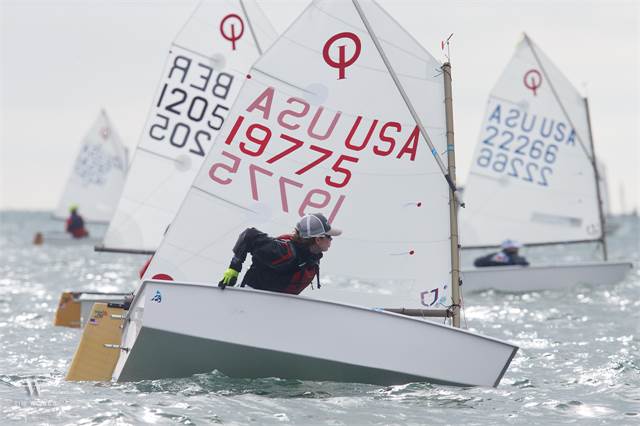 Young sailors compete in small sailboats on the water during a regatta. The boat in the foreground has 'USA 19775' on its sail, and others with similar numbers follow behind. The weather appears cloudy.