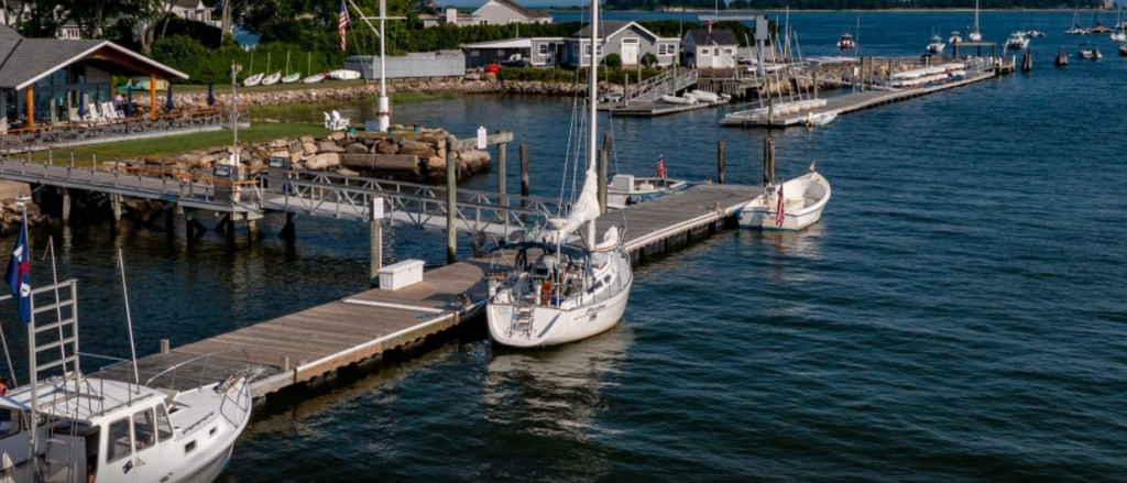 A marina with several docks and sailboats on the water, next to a grassy area and a building with outdoor seating. More boats are anchored in the background under a clear sky.
