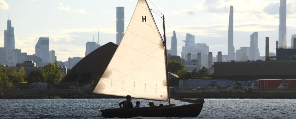 A small sailboat with three people aboard glides on the water, its sail glowing in sunlight. A city skyline with tall buildings is visible in the background under a partly cloudy sky.