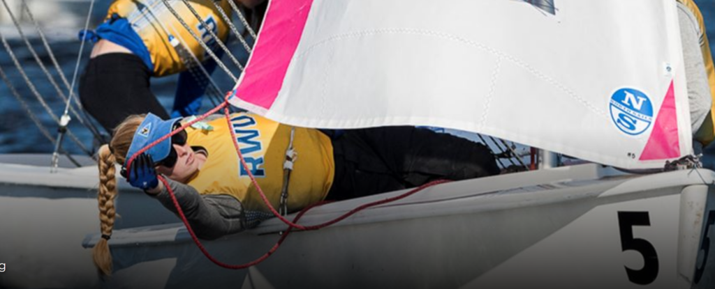 A sailor in a yellow life jacket and blue headband leans back over the side of a small sailboat, holding red ropes, as the white and pink sail partially obscures her face. The sailboat is marked with a number 5.