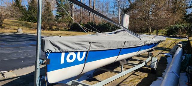 A blue and white sailboat with the number 180 covered by a gray tarp sits on a metal trailer next to a covered pool in an outdoor area with trees and grass.