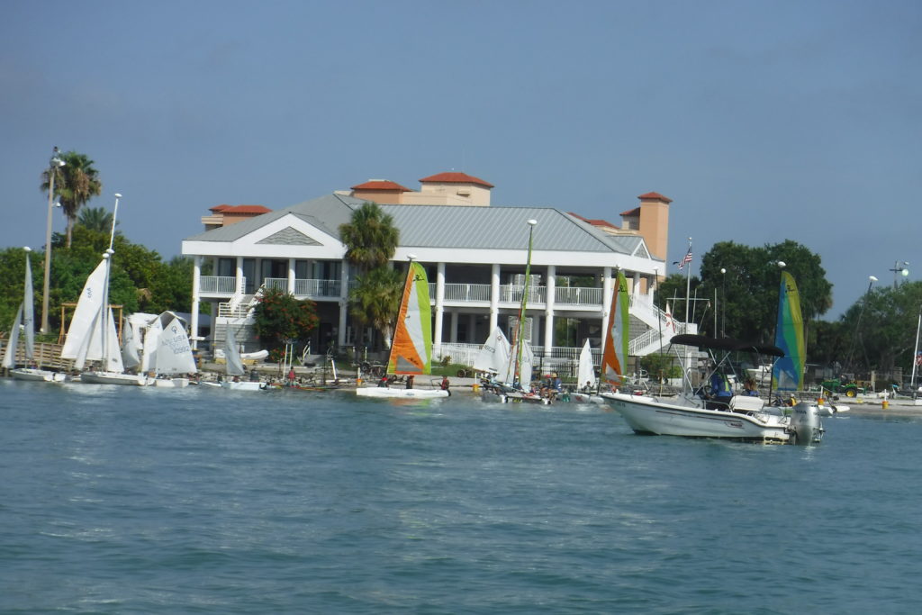 A waterfront building with red roofs stands behind a marina where several small sailboats and a motorboat are docked, with palm trees and greenery nearby under a partly cloudy sky.