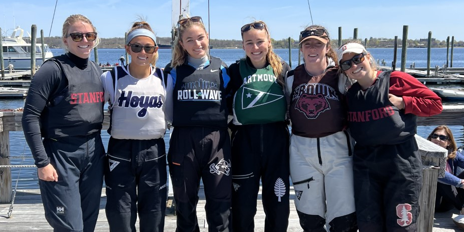 Six women sailors stand side by side on a dock, smiling and wearing sailing gear along with sweatshirts from different universities. Boats and water are visible in the background under a sunny sky.