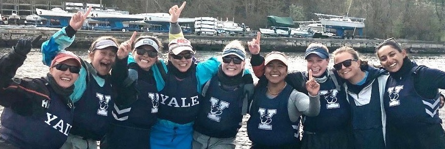 A group of nine women in matching Yale athletic gear stand on a dock by the water, smiling and holding up one finger, celebrating together with boats and trees in the background.