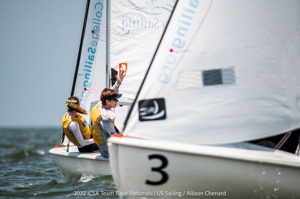 Two sailors compete in a small sailboat marked with the number 3 on the hull. One sailor raises an orange flag, while the other looks forward. The water is calm under a clear sky.