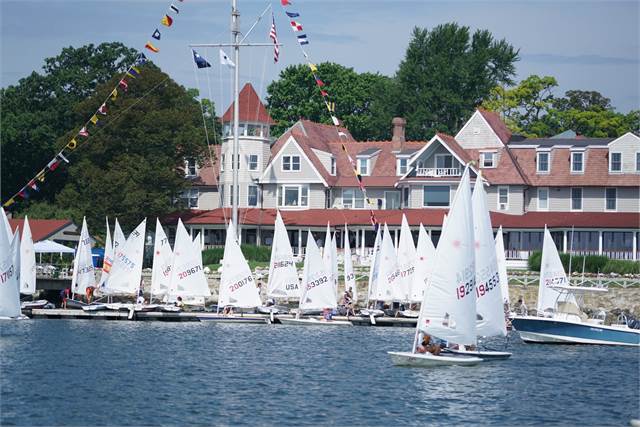 Several small sailboats with white sails gather near the shore in front of a large, Victorian-style building with red roofs and multiple flags flying. Trees and blue sky are in the background.