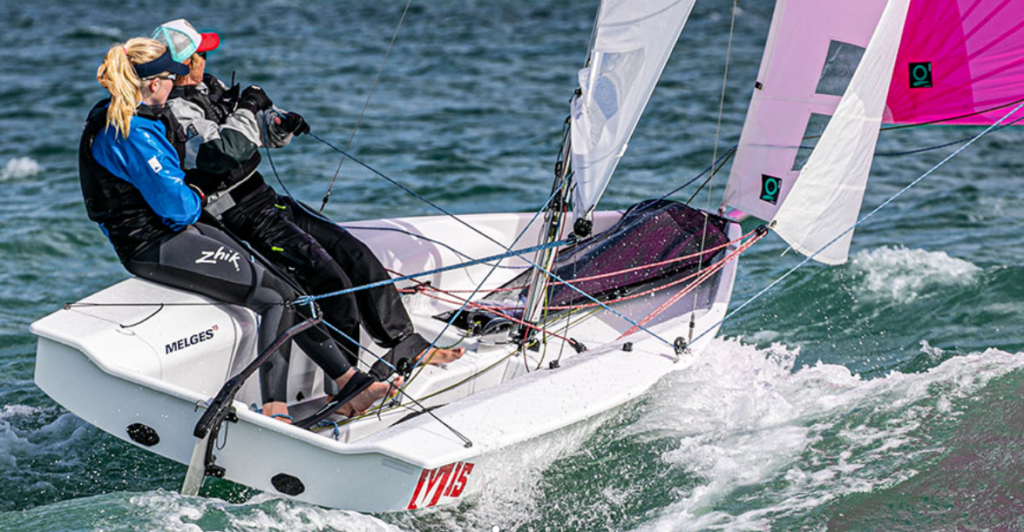 Two people sail a small white sailboat with pink and white sails on choppy water. Both wear sailing gear and lean to balance the boat as waves splash against the hull.