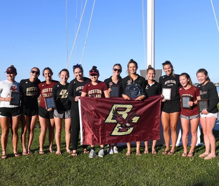 A group of thirteen women, some holding trophies and plaques, stand smiling outdoors on grass, holding a Boston College flag. They're dressed in Boston College sportswear, with a blue sky and flagpole in the background.