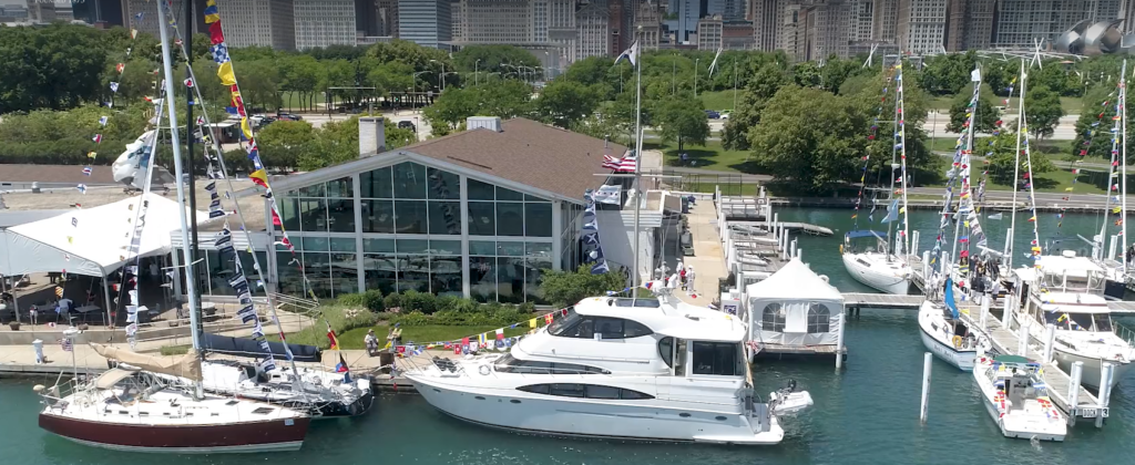 A marina with several boats decorated with colorful flags docked near a modern glass building, with trees and city buildings visible in the background.