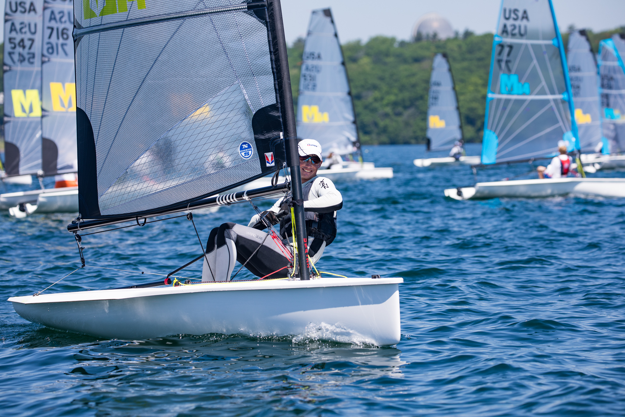 A person sailing a small racing boat on blue water, surrounded by other sailboats during a sunny day. Green trees and more boats are visible in the background. The main sailor is wearing sunglasses, a white cap, and a life jacket.