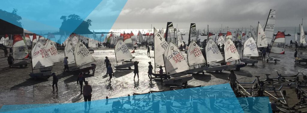 A group of people and numerous small sailboats with white sails gathered on a wet, sandy shore under a cloudy sky, with more boats visible on the water in the background. Blue graphic overlays are present.