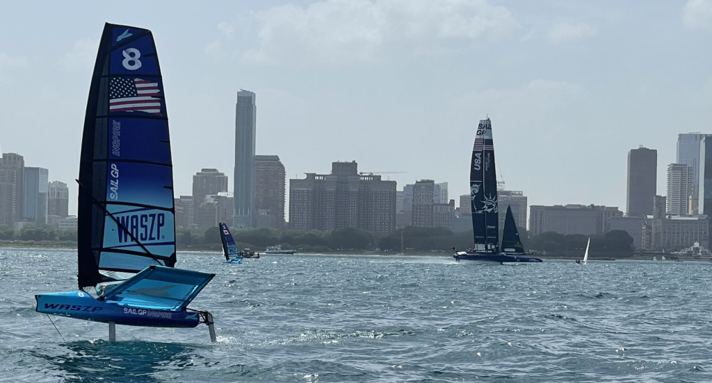 Two sailboats with hydrofoils race on choppy water near a city skyline on a hazy day. One sailboat in the foreground displays the number 8 and American flag, while tall buildings rise in the background.