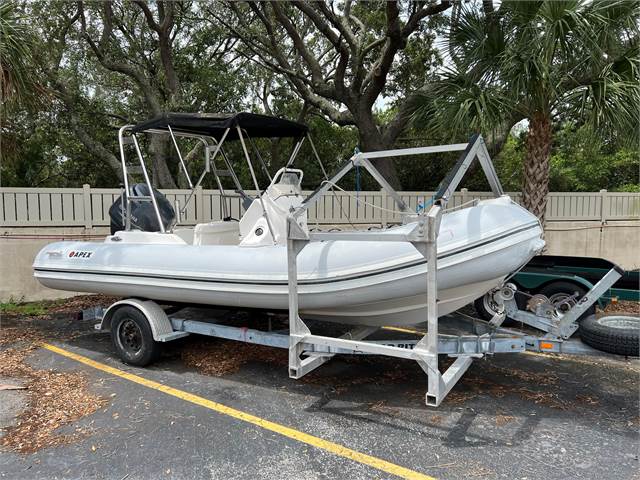 A white inflatable boat with a black canopy is secured on a metal trailer in a parking lot, surrounded by trees and a tan fence.