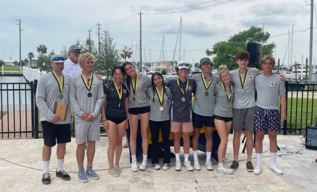 A group of ten young people and two adults stand outdoors by a marina, all wearing medals and matching gray shirts. Some hold plaques, and everyone is smiling. Sailboats and trees are visible in the background.