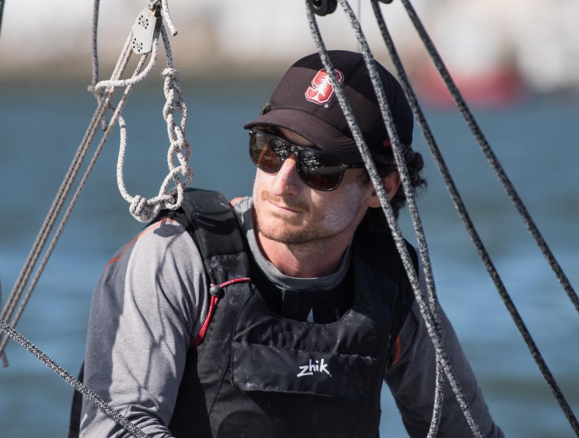 A man wearing sunglasses, a black cap, and a life vest sits on a sailboat, surrounded by ropes, with water and an out-of-focus shoreline in the background.