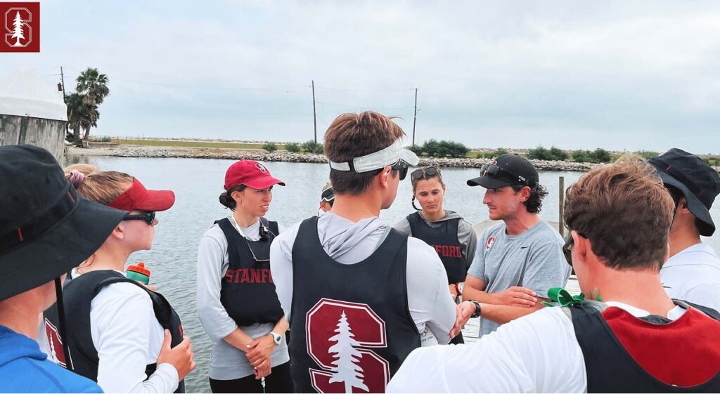 A group of Stanford rowers in uniforms stand together outdoors near water, listening to a coach who is gesturing during a team meeting or briefing. It is daytime with cloudy skies.