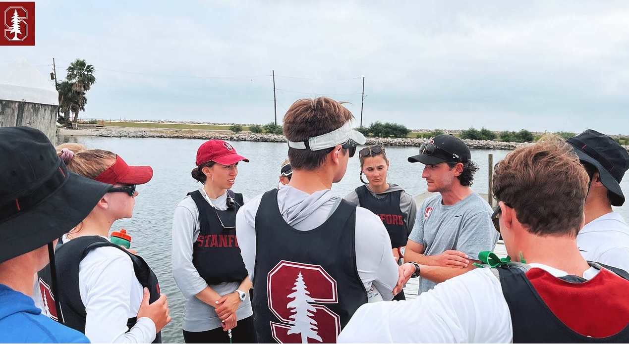 A group of Stanford rowers in uniforms stand together outdoors near water, listening to a coach who is gesturing during a team meeting or briefing. It is daytime with cloudy skies.