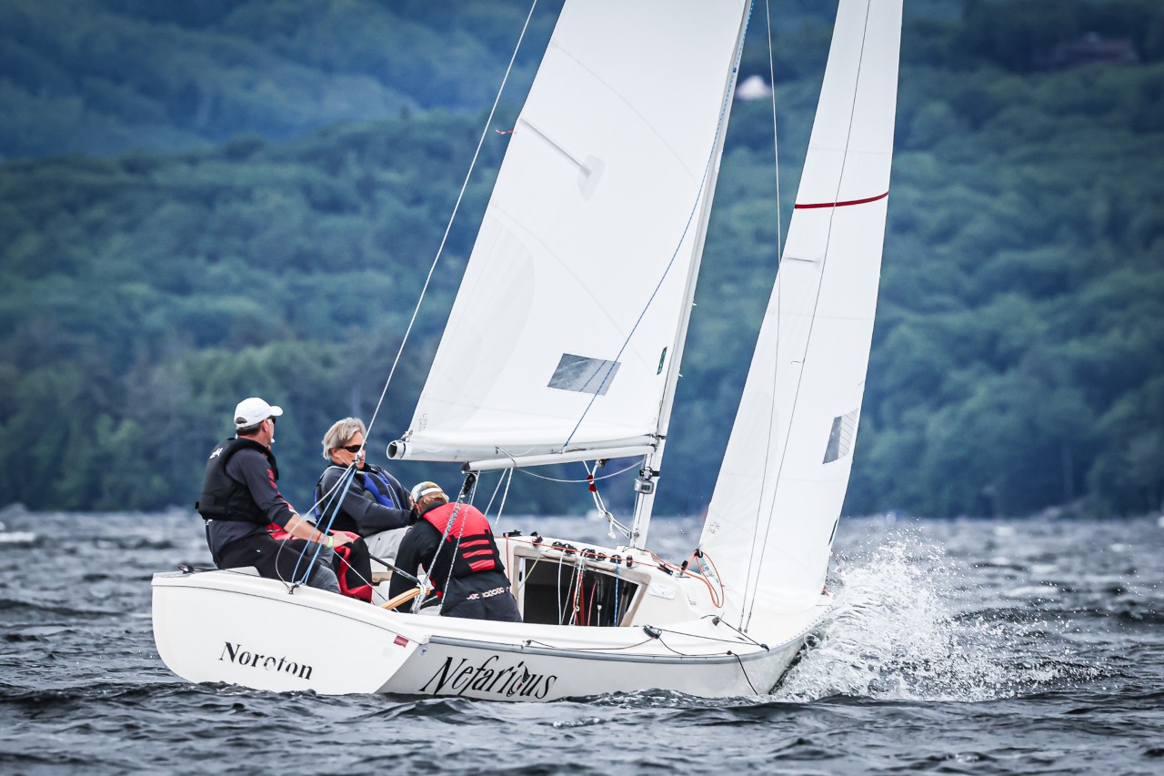 Three people in life jackets sail a small white sailboat named "Nefarious" on choppy water, with green forested hills in the background. Spray splashes at the bow as they navigate.