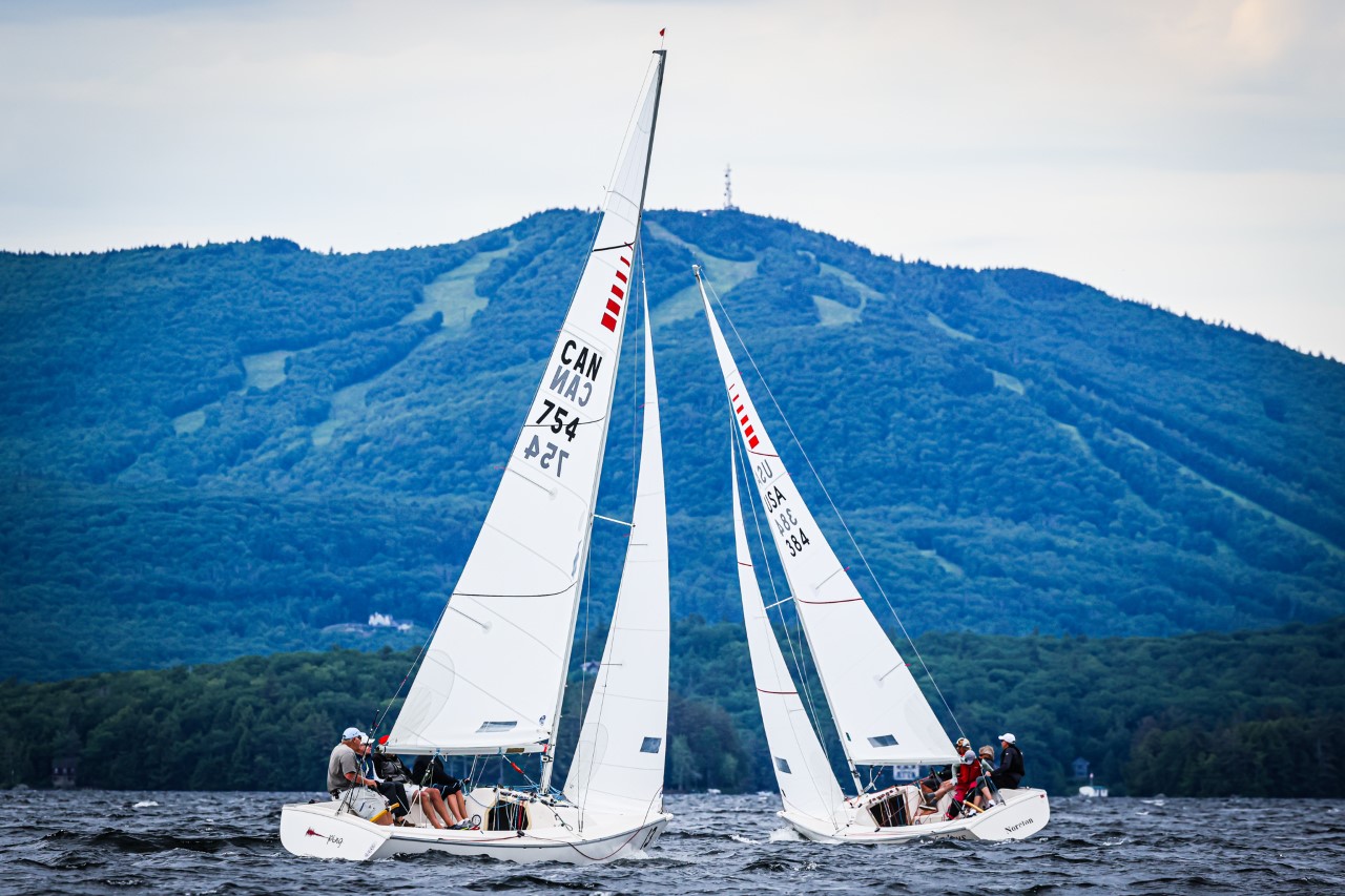Two white sailboats with crews are racing on a lake, with forested hills and a mountain in the background under a cloudy sky.