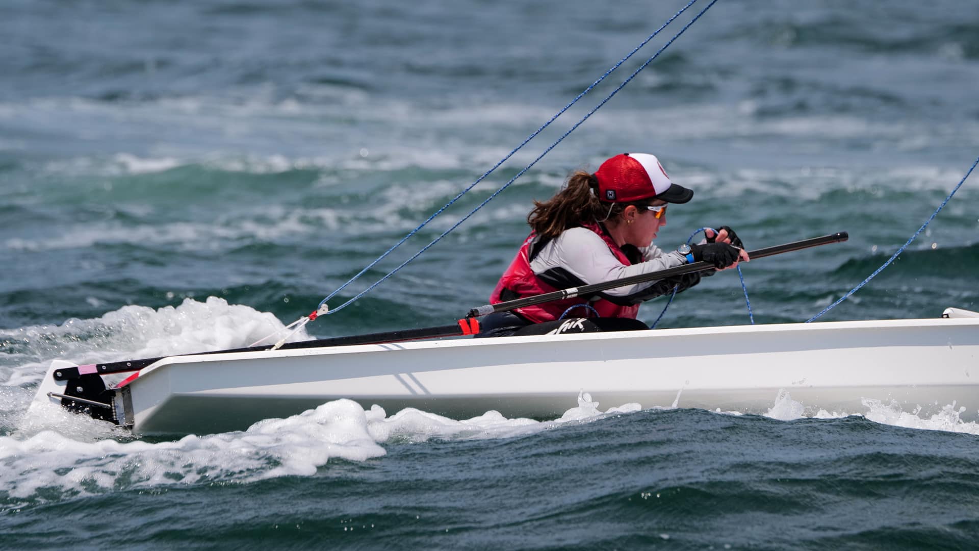 A person wearing a red vest and cap sails a small white boat through choppy blue water, gripping the tiller and looking ahead with focus.