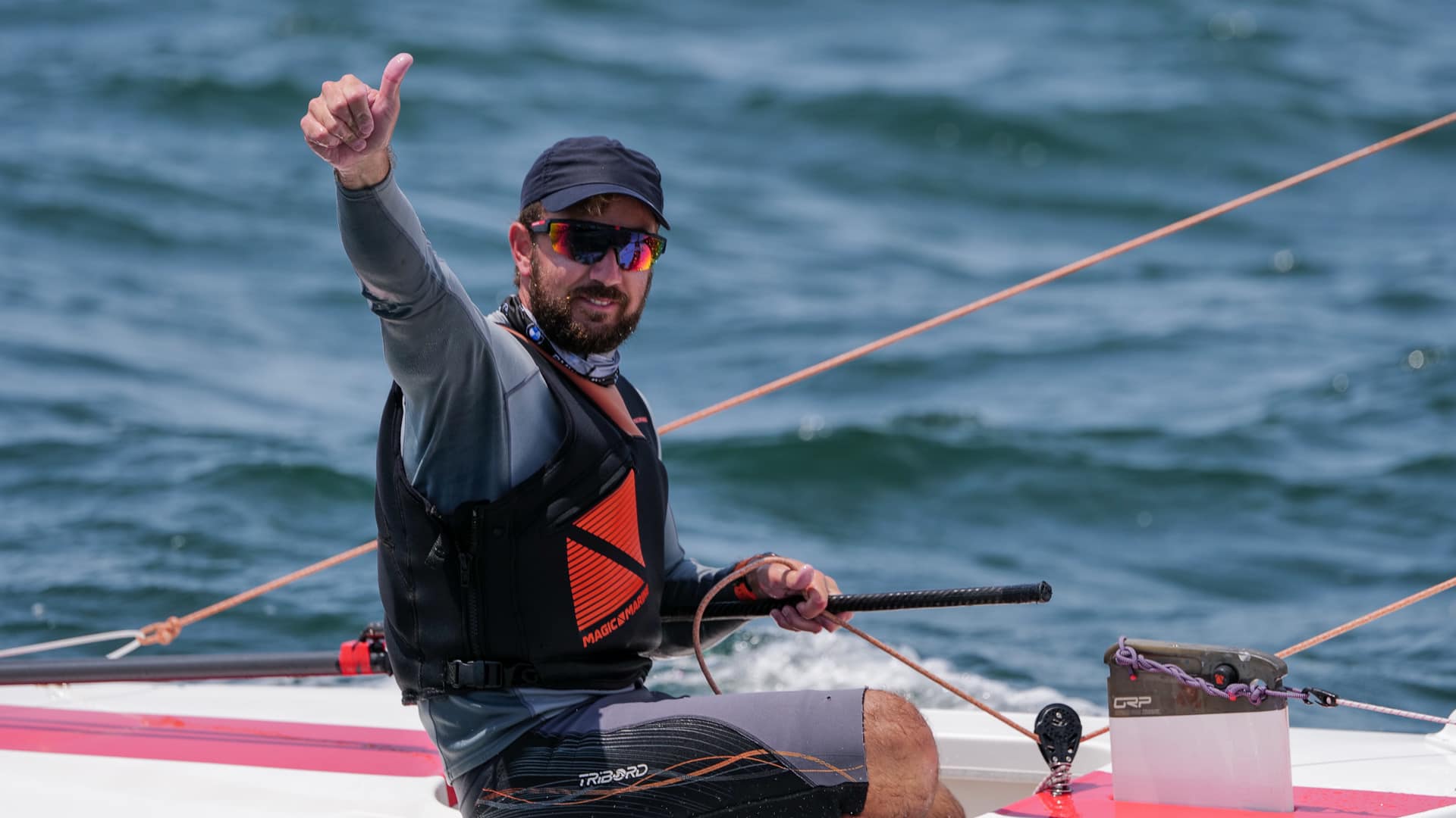 A man wearing sunglasses, a cap, and a life vest gives a thumbs-up while sailing a small boat on the water.