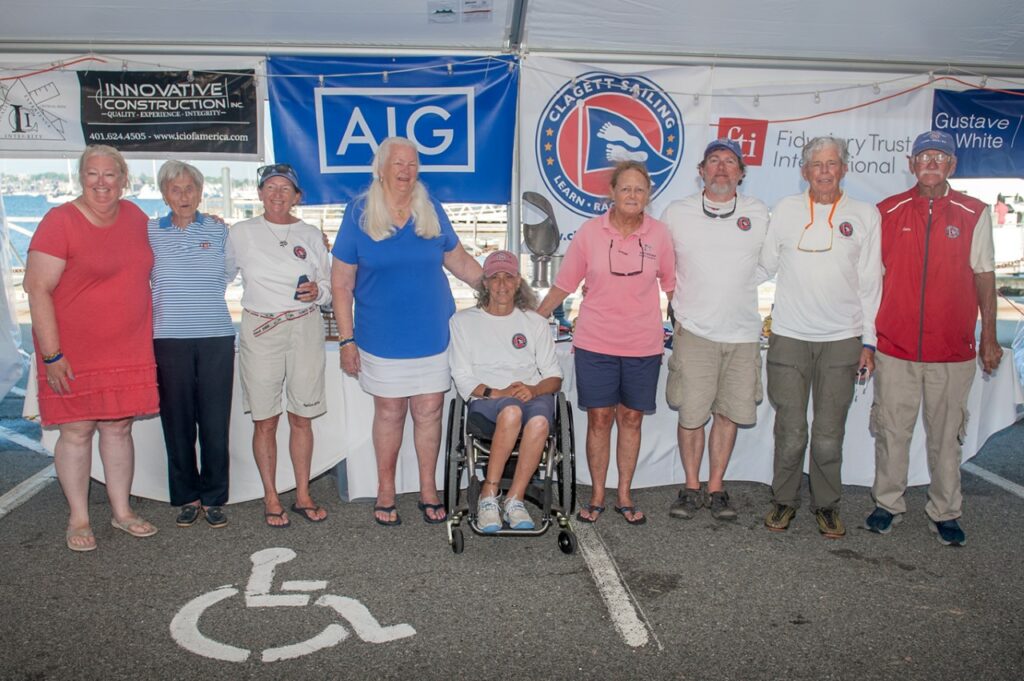 Ten people, some in matching white shirts and caps, stand and smile under a tent with banners behind them. One person is seated in a wheelchair. They are on a paved area next to a marina.