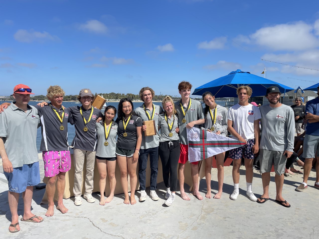 A group of ten young people, wearing medals and casual clothes, stand barefoot on a sunny dock with two adult men. Some hold a small flag and a plaque; blue umbrellas and water are in the background.