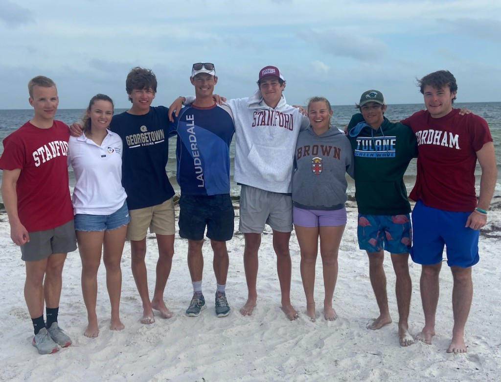 Eight young adults stand barefoot on a sandy beach, arm in arm, smiling at the camera. They wear T-shirts and sweatshirts from various universities. The ocean and cloudy sky are in the background.